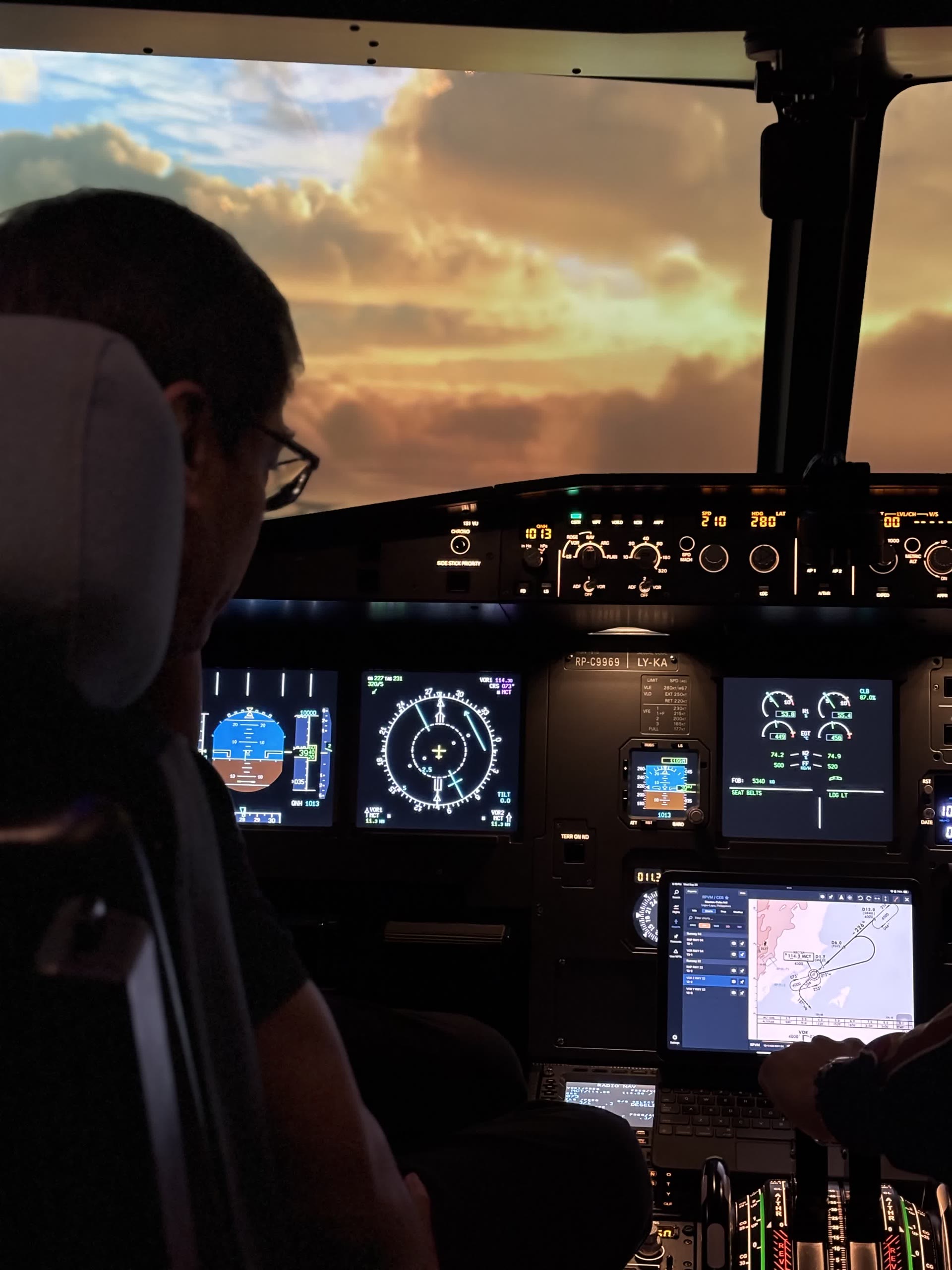 A pilot seated at the controls of the A320 fixed-base simulator, all instrument displays lit, with a dramatic sunset cloudscape shown on the outside visual.